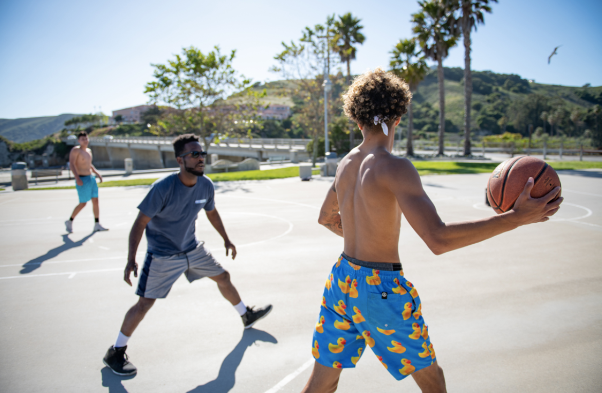 A small group of young mean are playing basketball outside with palm trees in the background. The man in the foreground is shirtless and handles the basketball. 