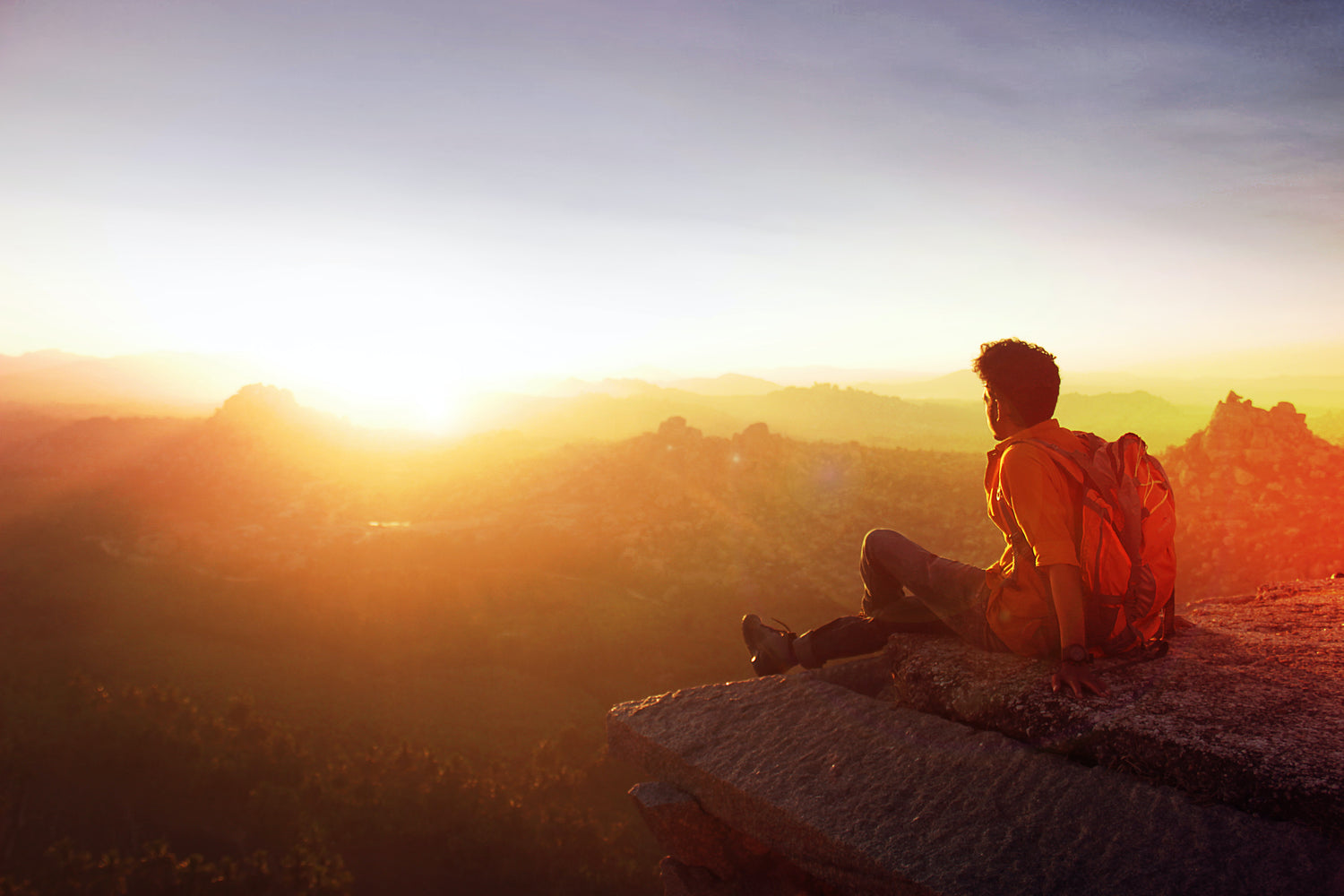 A man sits on a cliff looking at the sunrise over the mountains. 