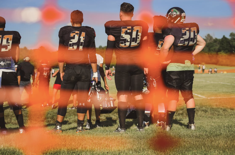 A ground of high school football players standing in a group between exercises photographed from behind an orange fence for effect. 