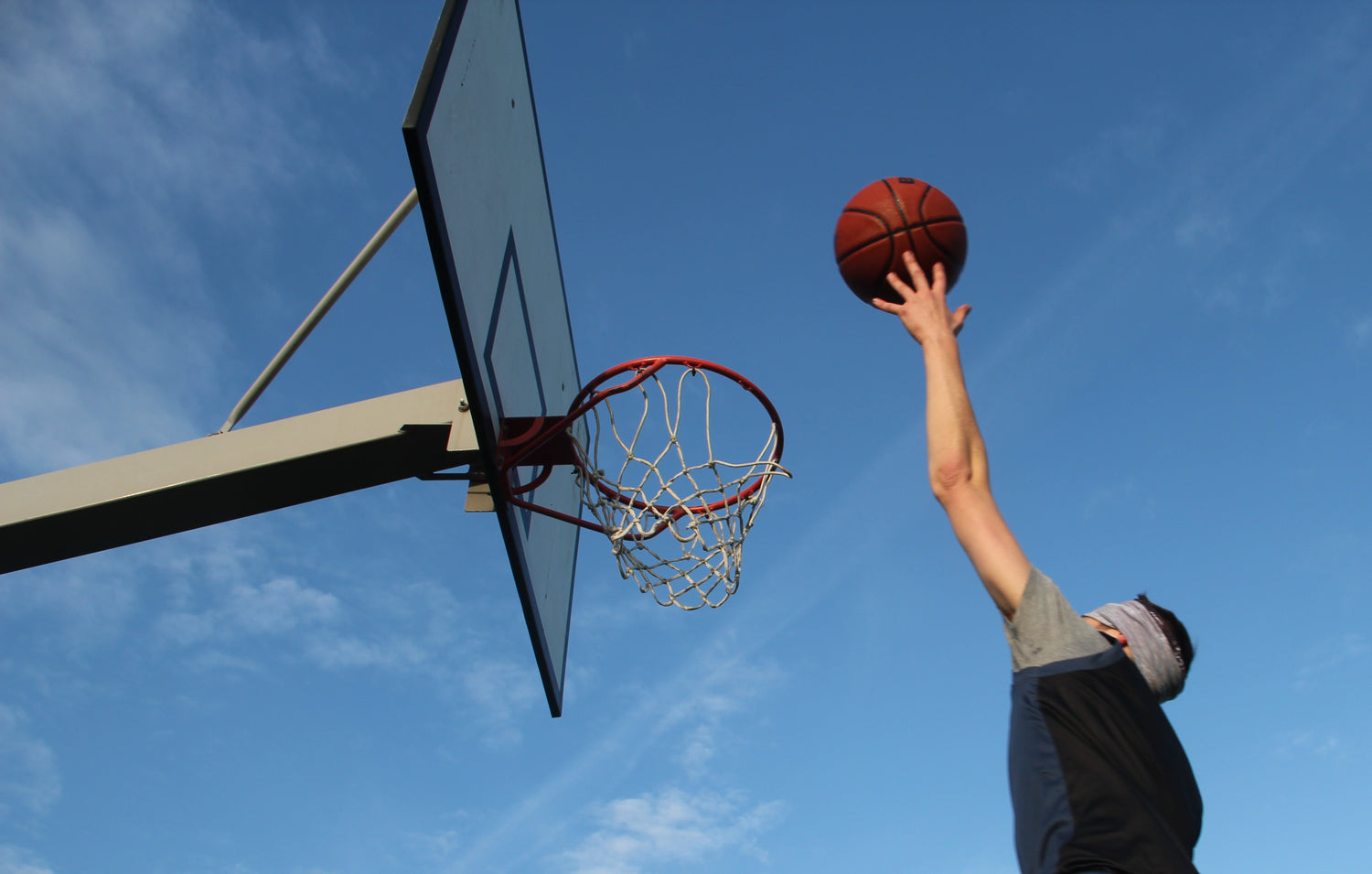 photo of a man doing a lay up at a basketball hoop. the photo is taken from the ground looking up with a clear blue sky. 